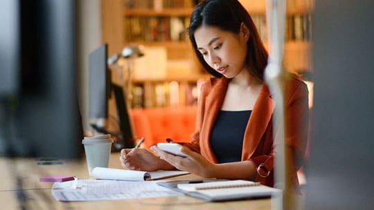 Woman seated at desk looks at a calculator looks at a calculator in her left hand.